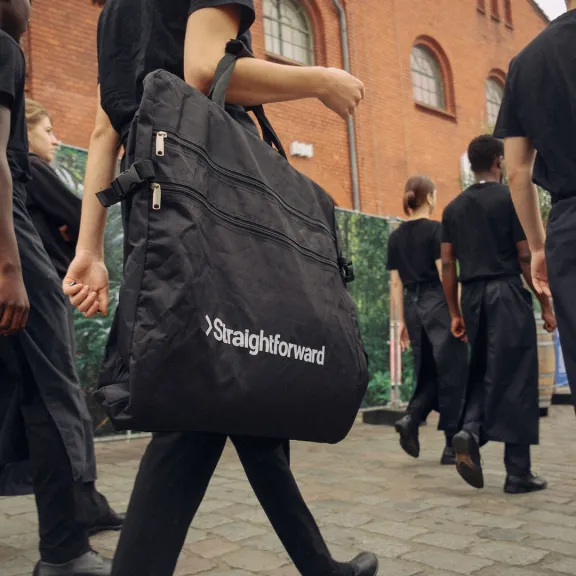 Mehrere Personen in schwarzer Kleidung gehen auf gepflastertem Weg vor roten Backsteingebäuden.