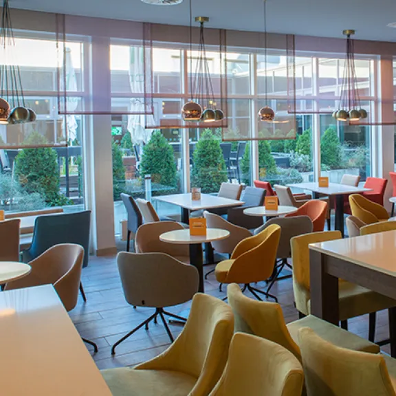 Modernes Café mit bunten Stühlen, runden Tischen und großen Fenstern mit Blick nach draußen.