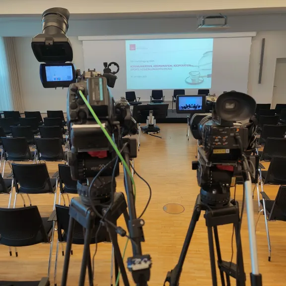 Leerer Konferenzraum mit zwei Kameras, Stuhlreihen und Bühne mit Projektionsleinwand.