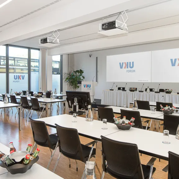 Heller Konferenzraum mit Tischen, Stühlen, Getränken und zwei großen Leinwänden.