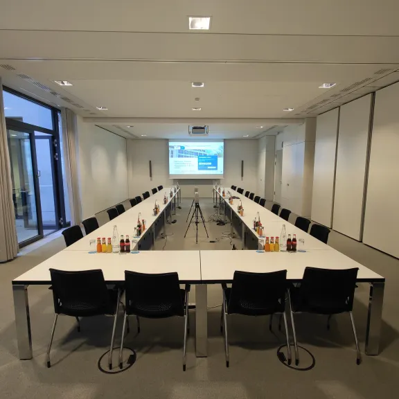Konferenzraum mit U-förmig angeordneten Tischen, Stühlen und Beamer-Leinwand