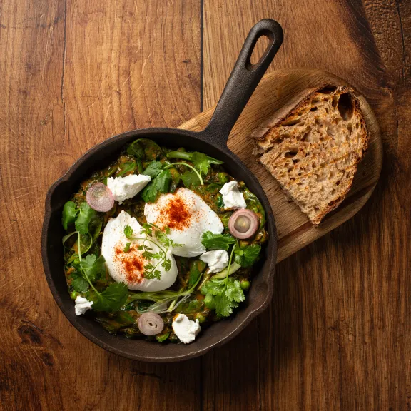Pfanne mit pochierten Eiern, Spinat und Brot auf Holzunterlage.