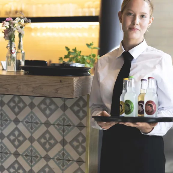Kellnerin mit Tablett und drei Getränkeflaschen steht an einem Tresen.