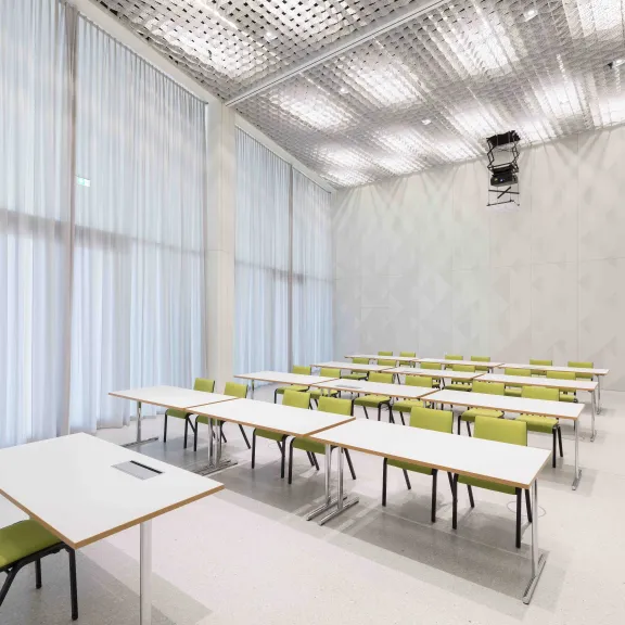 Heller Seminarraum mit weißen Tischen und grünen Stühlen, große Fenster mit Vorhängen.