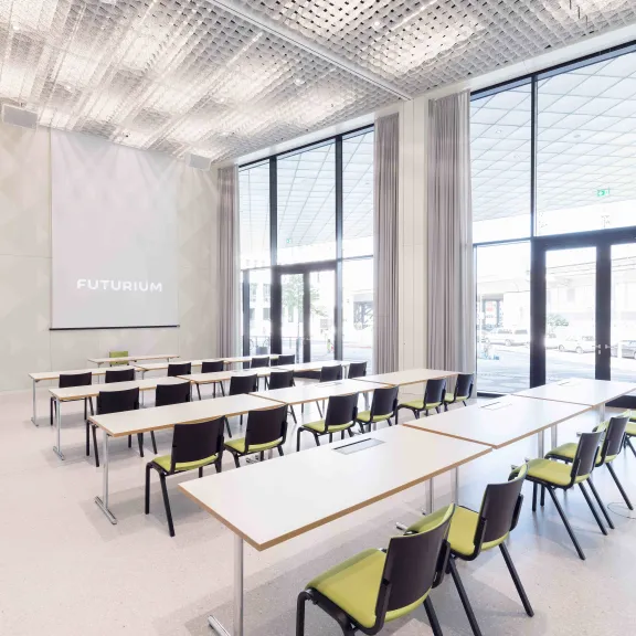 Heller, leerer Seminarraum mit Tischen, Stühlen und großen Fenstern.