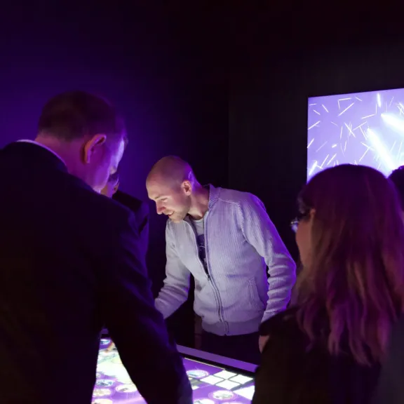 Mehrere Personen interagieren mit einem leuchtenden Touchscreen-Tisch in dunklem Raum.