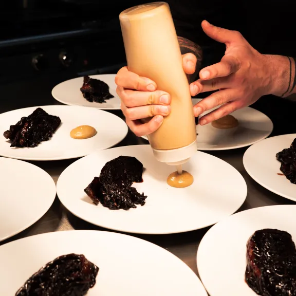 Koch gibt Sauce auf mehrere Teller mit dunklem Fleischgericht.