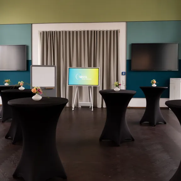 Konferenzraum mit Stehtischen, Blumenvasen, Monitoren und Flipchart.