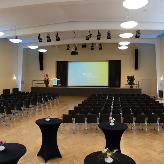 Heller Konferenzsaal mit Bühne, Stuhlreihen und Stehtischen, große Leinwand vorne.