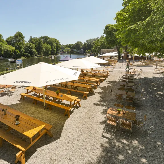Sunny beer garden with wooden tables by the river, large parasols, trees providing shade.