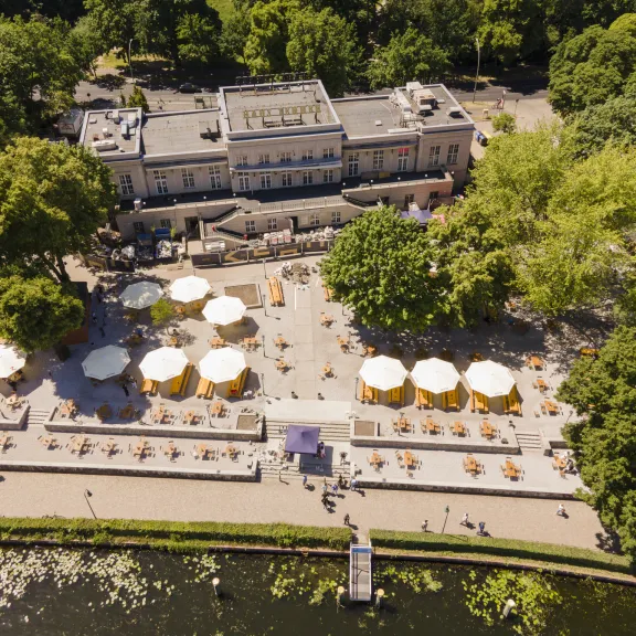 Luftaufnahme eines Cafés mit Sonnenschirmen und Tischen im Park am Wasser.