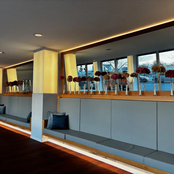 Moderner Warteraum mit langen Sitzbänken, Kissen und Vasen mit Blumen am Fenster.