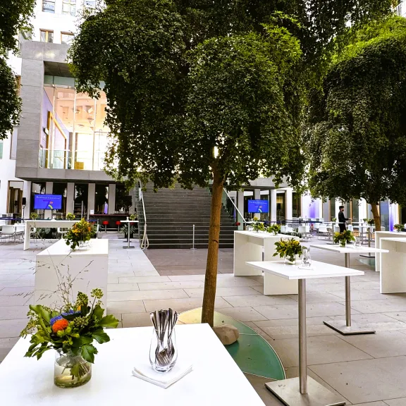 Moderner Innenhof mit weißen Tischen, Blumendekorationen und großen grünen Bäumen.