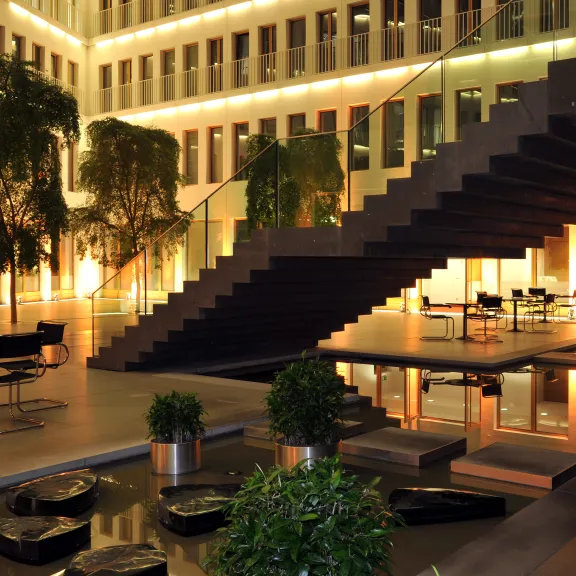 Modernes, beleuchtetes Atrium mit Sitzbereichen, Pflanzen und einer breiten Treppe.