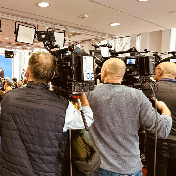 Pressekonferenz mit Kameraleuten, Fotografen und Publikum in einem hellen Raum.
