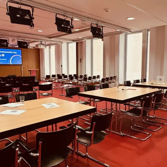 Konferenzraum mit roten Teppich, leeren Tischen und Stühlen, Präsentationsbildschirm vorne.