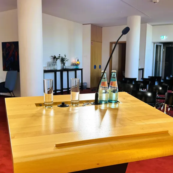 Rednerpult mit Mikrofon und Wasserflaschen vor leeren Stuhlreihen in Konferenzraum.