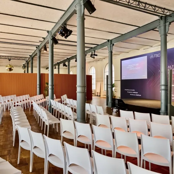 Konferenzraum mit weißen Stühlen und großer Bühne, zwei Bildschirme, leerer Saal.