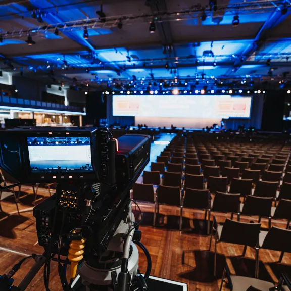 Kamera filmt leeren Konferenzsaal mit beleuchteter Bühne im Hintergrund.