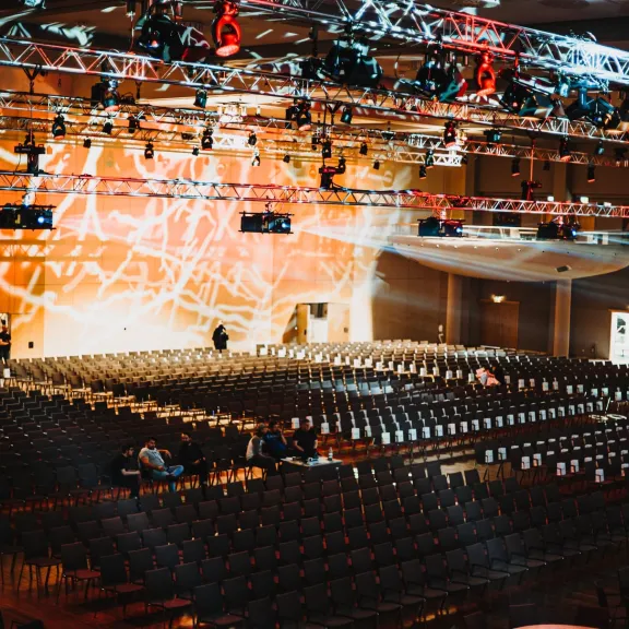 Großer Veranstaltungssaal mit vielen leeren Stuhlreihen und warmer Beleuchtung.