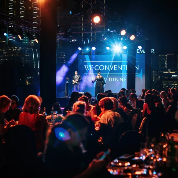 Menschen sitzen an runden Tischen bei einer Gala mit beleuchteter Bühne im Hintergrund.