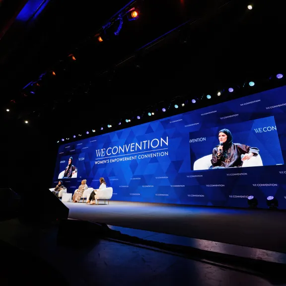 Das Bild ist dunkel. Zu sehen ist rechts im Hintergrund eine LED Wand, welche einen blauen Hintergrund zeigt, weiße Schrift drauf hat und zwei Bilder im Bild. Auf den Bildern zu sehen ist eine der drei Sprecherinnen, die vor der LED-Wand auf Sesseln sitzen.