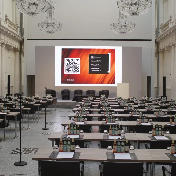 Großer Konferenzraum mit vielen Tischen, Stühlen und eine Leinwand mit Projektion vorne.