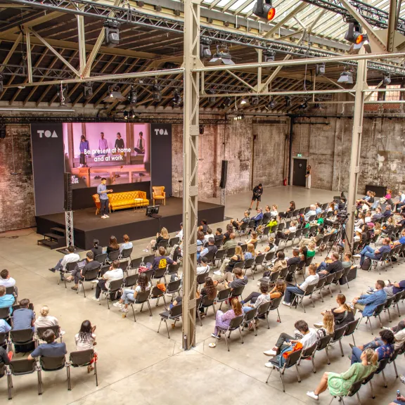 Zu sehen ist eine Fabrikhalle. Links gehalten im Hintergrund ist eine kleine Bühne, mit LED Wand, einem Sofa , ein Stuhl und einer Person, die darauf stehen.  Vor der Bühne sind Stühle in einigen Reihen aufgestellt und sie sind fast alle voll besetzt.