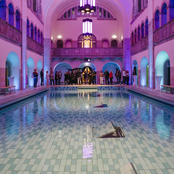 Beleuchtetes Hallenbad mit lila Licht, Menschen stehen am Beckenrand.