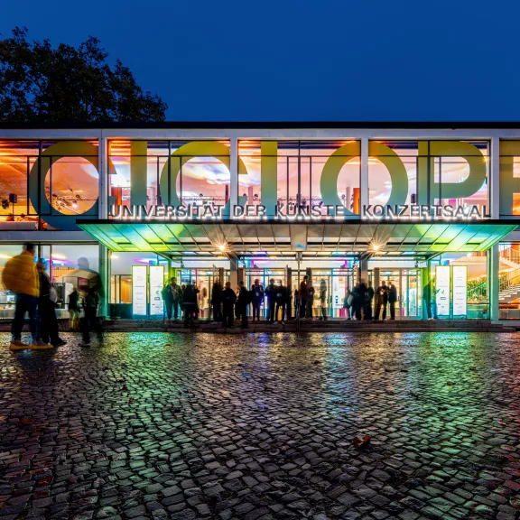 Modernes, bunt beleuchtetes Gebäude bei Nacht mit Menschen vor dem Eingang.