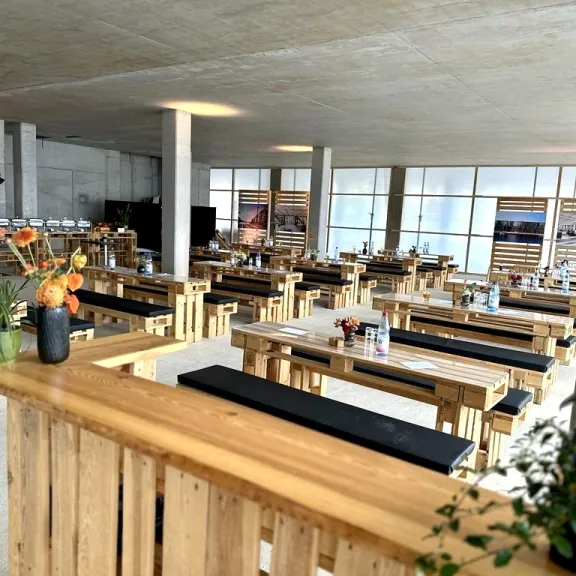 Heller Raum mit Holzbänken und Tischen, Blumenvasen auf den Tischen.