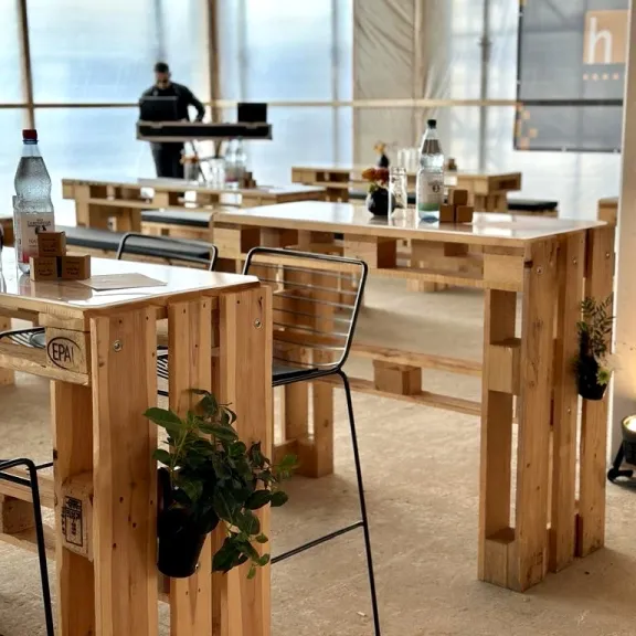 Heller Raum mit Tischen aus Paletten, Pflanzen, Getränkeflaschen und einer Person im Hintergrund.