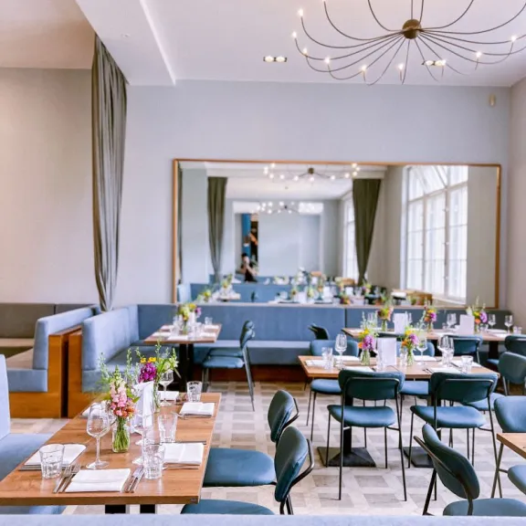 Modernes Restaurant mit blauen Stühlen, Holztischen und Blumenvasen, heller Raum.