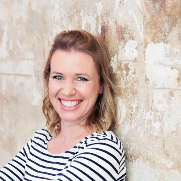 Frau mit blondem Haar lächelt vor einer hellen, strukturierten Wand.