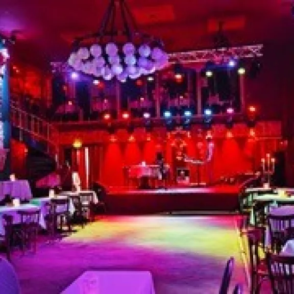 Stimmungsvoll beleuchteter Saal mit runden Tischen, Bühne im Hintergrund, rotes Licht.