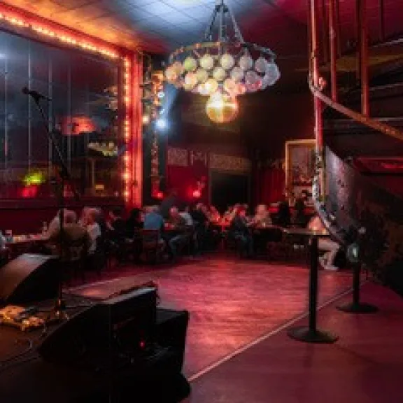 Gemütlicher Club mit rotem Licht, Bühne und sitzenden Gästen an Tischen.