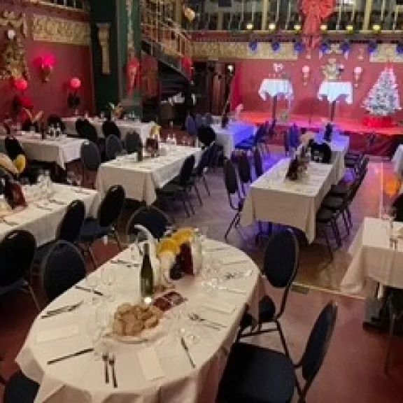 Festlich gedeckter Saal mit runden Tischen, Bühne und beleuchtetem Weihnachtsbaum.