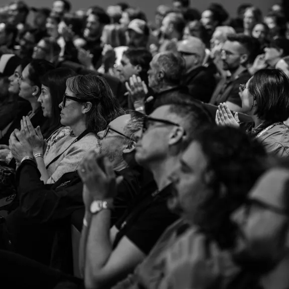 Schwarzweißfoto mehrerer applaudierender Menschen in einem voll besetzten Saal.