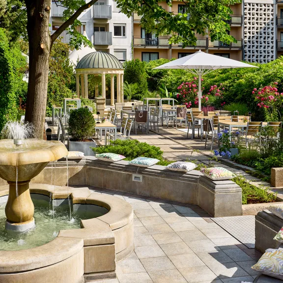 Sonniger Garten mit Springbrunnen, Tischen, Stühlen und vielen Pflanzen.