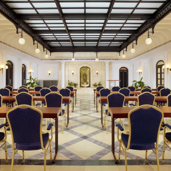 Eleganter Konferenzraum mit Reihen blauer Stühle und Tischen, hohe Decke, helle Beleuchtung.