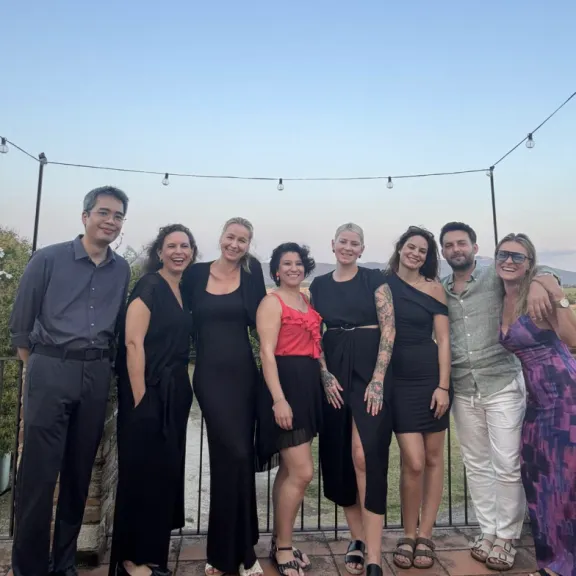 Acht Personen posieren lächelnd auf einer Terrasse bei Sonnenuntergang.