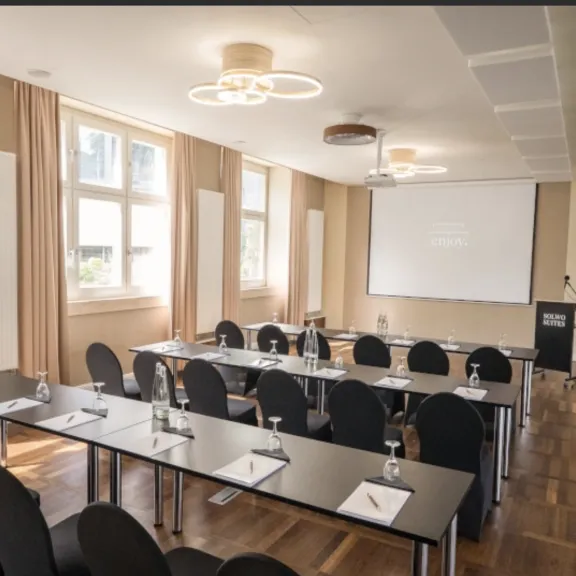 Heller Konferenzraum mit Tischen, Stühlen, Schreibmaterial und Projektionsleinwand.