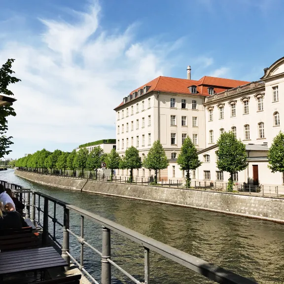Sonniger Fluss mit Uferpromenade, Bäumen und großem weißen Gebäude mit rotem Dach.