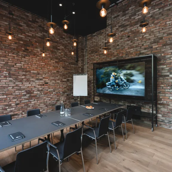 Moderner Konferenzraum mit langem Tisch, Stühlen und großem Bildschirm vor Backsteinwand.