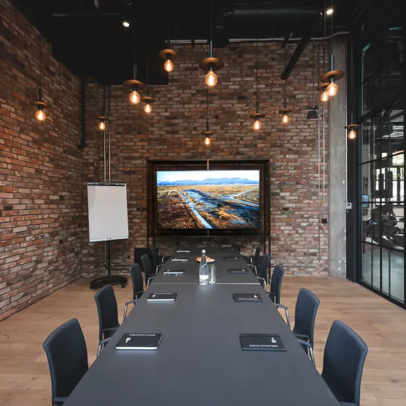 Moderner Konferenzraum mit langem Tisch, Stühlen und großem Bildschirm an Ziegelwand.
