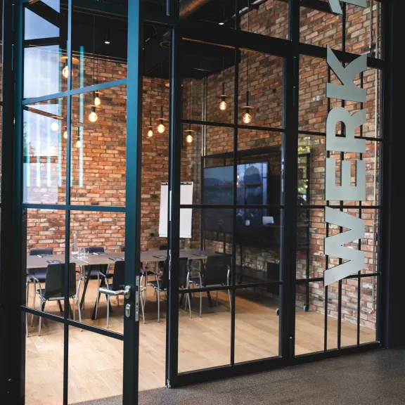 Glasraum mit Schriftzug „WERK 1“, Backsteinwand, Konferenztisch und Stühlen.