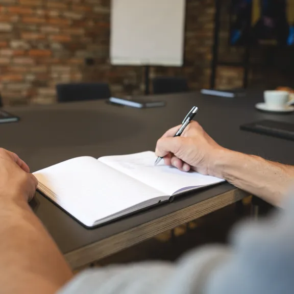 Person schreibt Notizen in ein aufgeschlagenes Notizbuch bei einer Besprechung.