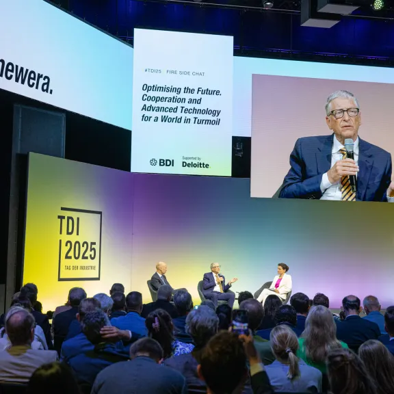 Podiumsdiskussion mit drei Personen vor großem Publikum, Bühne mit farbigem Licht.