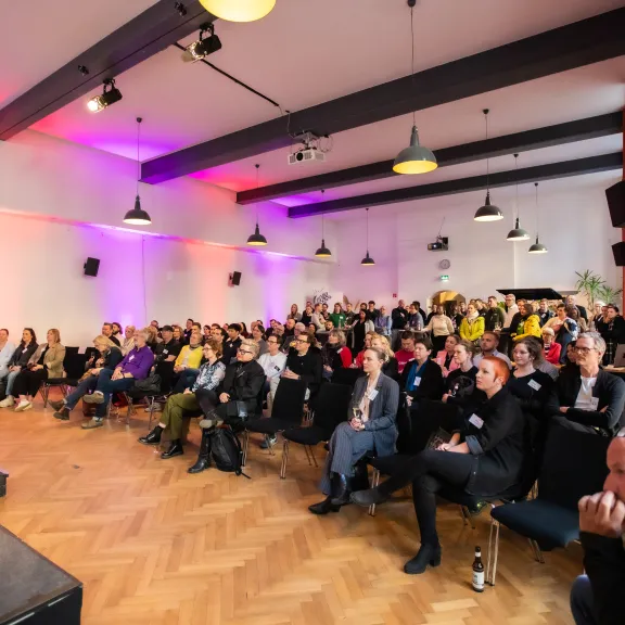 Publikum in einem bunt beleuchteten Konferenzraum, aufmerksam auf die Bühne gerichtet.
