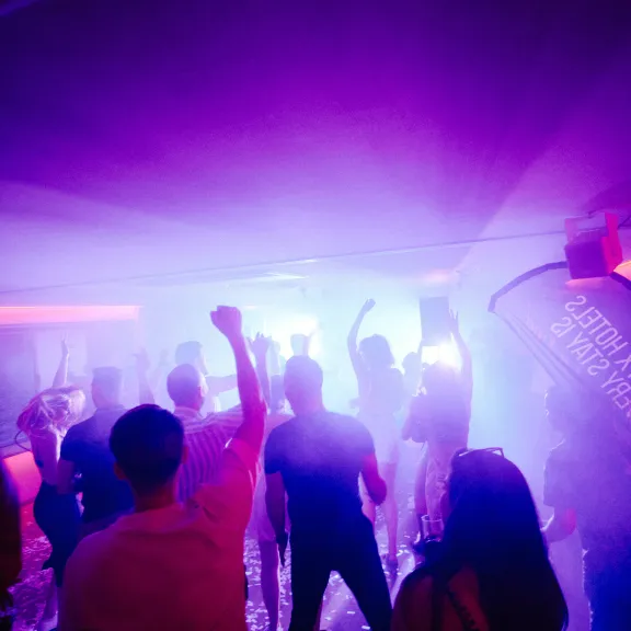 Menschen tanzen in einem Club mit lila Licht und Nebel.